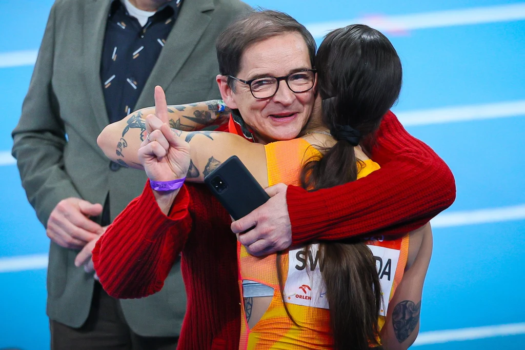 Mężczyzna w czerwonym swetrze obejmuje zawodniczkę na bieżni, oboje wyrażają radość; sportowczyni ma numer startowy oraz długie ciemne włosy.