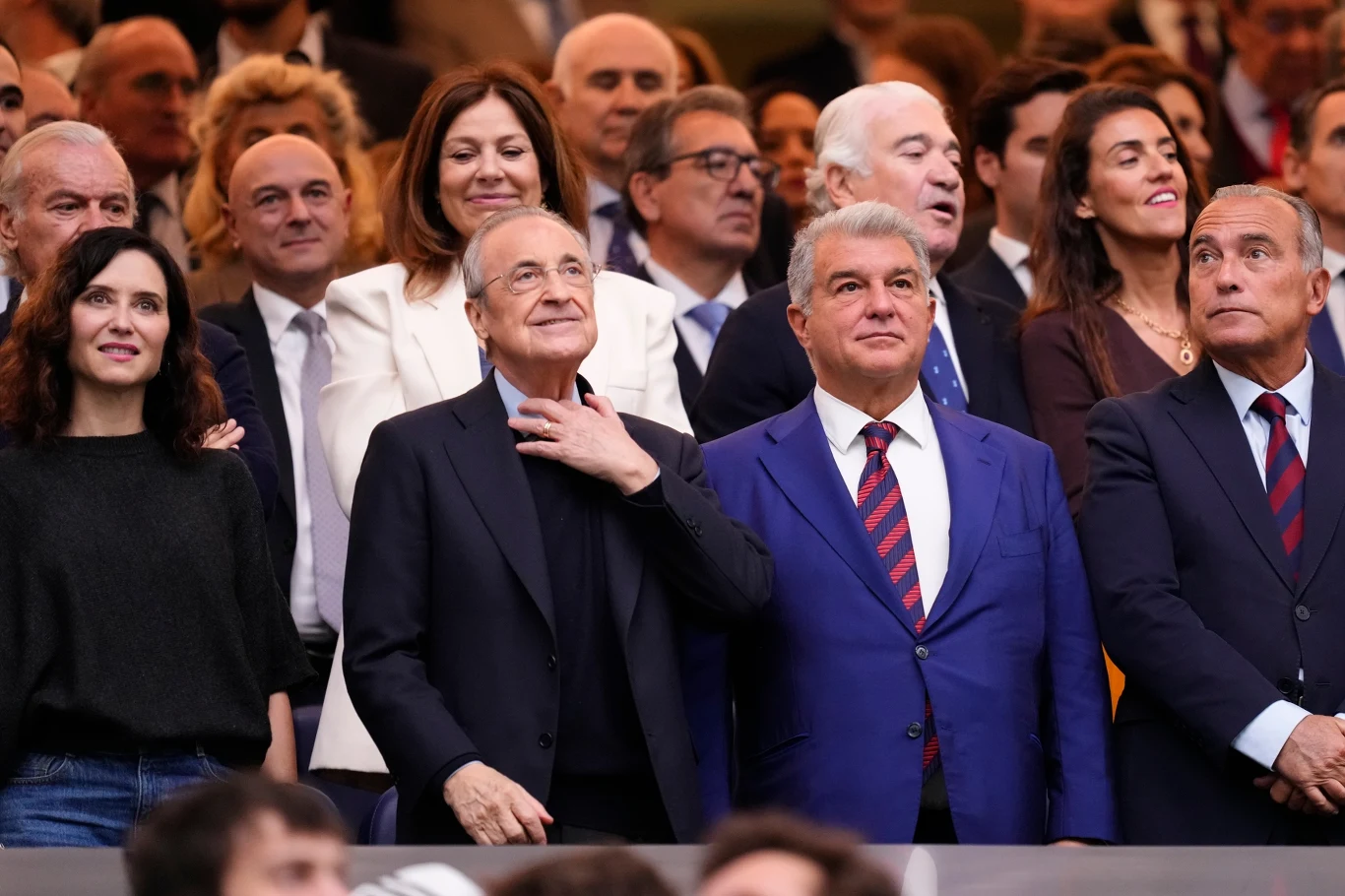 Florentino Perez i Joan Laporta Grupa elegancko ubranych osób na trybunach stadionu, skupionych na wydarzeniu sportowym, mężczyźni w garniturach, kobiety w formalnych strojach, radosna atmosfera, skupienie na kilku głównych postaciach w centrum kadru.
