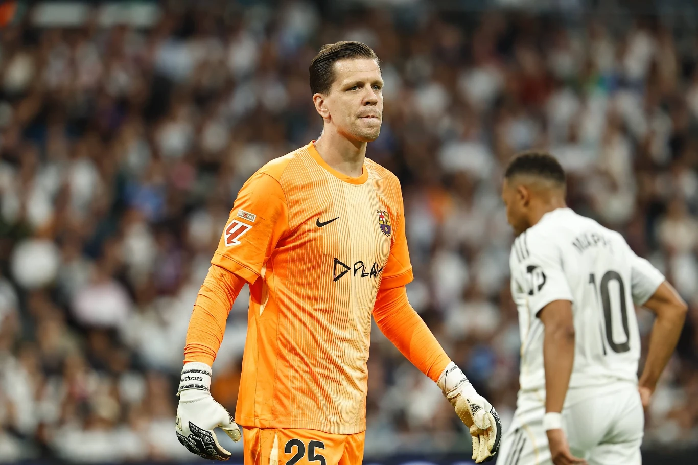 Bramkarz w pomarańczowym stroju sportowym stoi na boisku piłkarskim, za nim zawodnik drużyny przeciwnej w białej koszulce. W tle rozmyty tłum kibiców na trybunach.