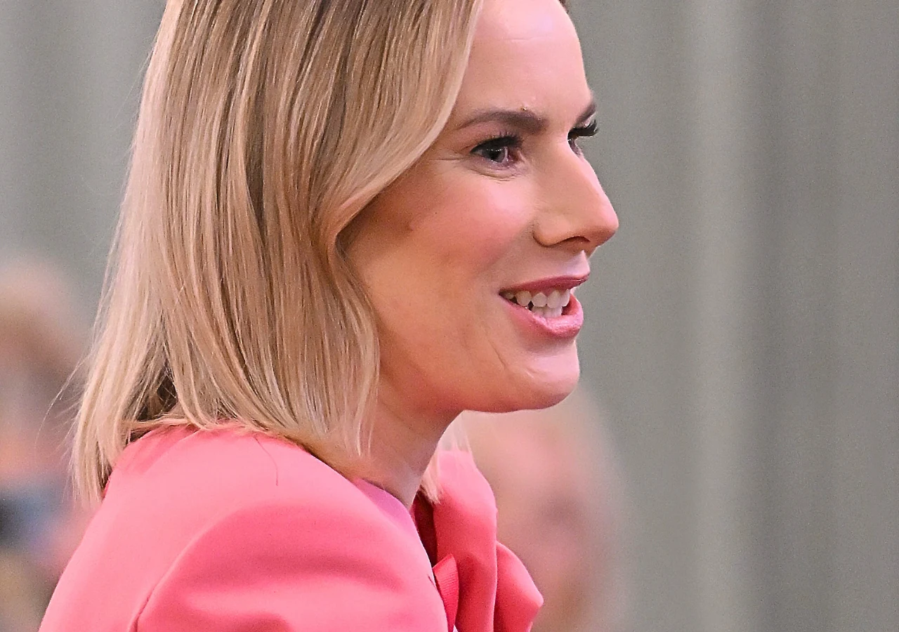 Marta Nawrocka Uśmiechnięta kobieta w jasnoróżowym żakiecie na tle rozmytego, jasnego wnętrza, włosy blond opadające na ramiona, twarz lekko zwrócona w bok.