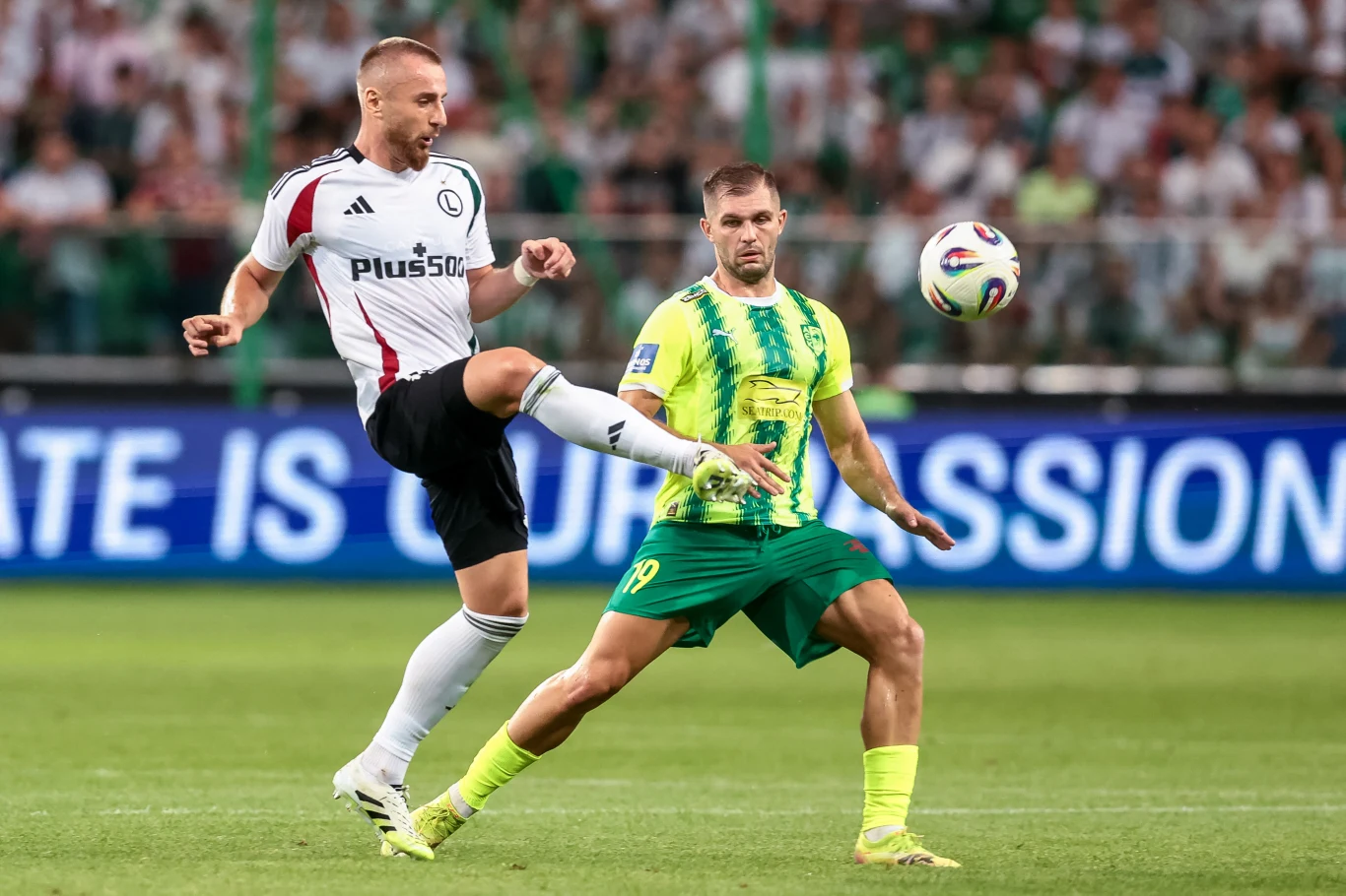 Karol Angielski strzelił jedną z bramek w dwumeczu AEK-u Larnaka z Legią Warszawa Dwóch piłkarzy walczących o piłkę na zielonym boisku podczas meczu piłkarskiego, trybuny wypełnione kibicami w tle.