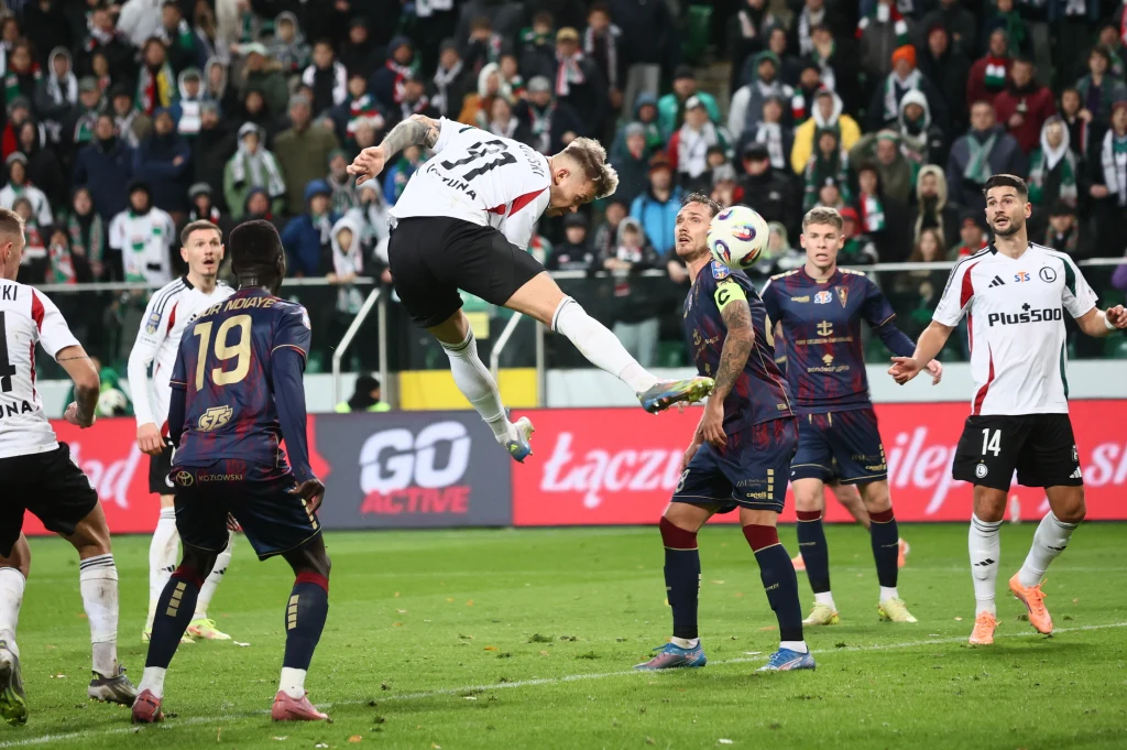 Kamil Piątkowski Zawodnik drużyny w białym stroju wyskakuje do główki przy bramce przeciwników, wokół zgromadzeni są gracze obu drużyn oraz sędzia boczny obserwujący akcję, w tle kibice na trybunach.