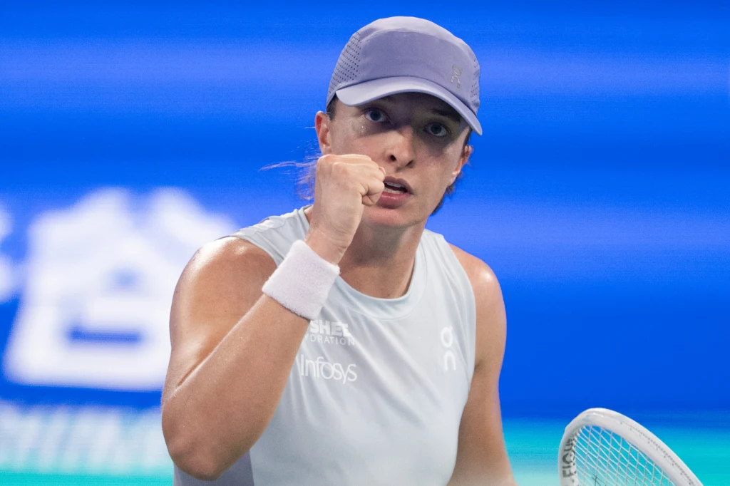 Iga Świątek Sportowiec w jasnej koszulce i czapce z daszkiem świętuje sukces na korcie tenisowym, wykonując gest zaciśniętej pięści. W tle rozmyte niebieskie tło i fragment rakiety tenisowej.