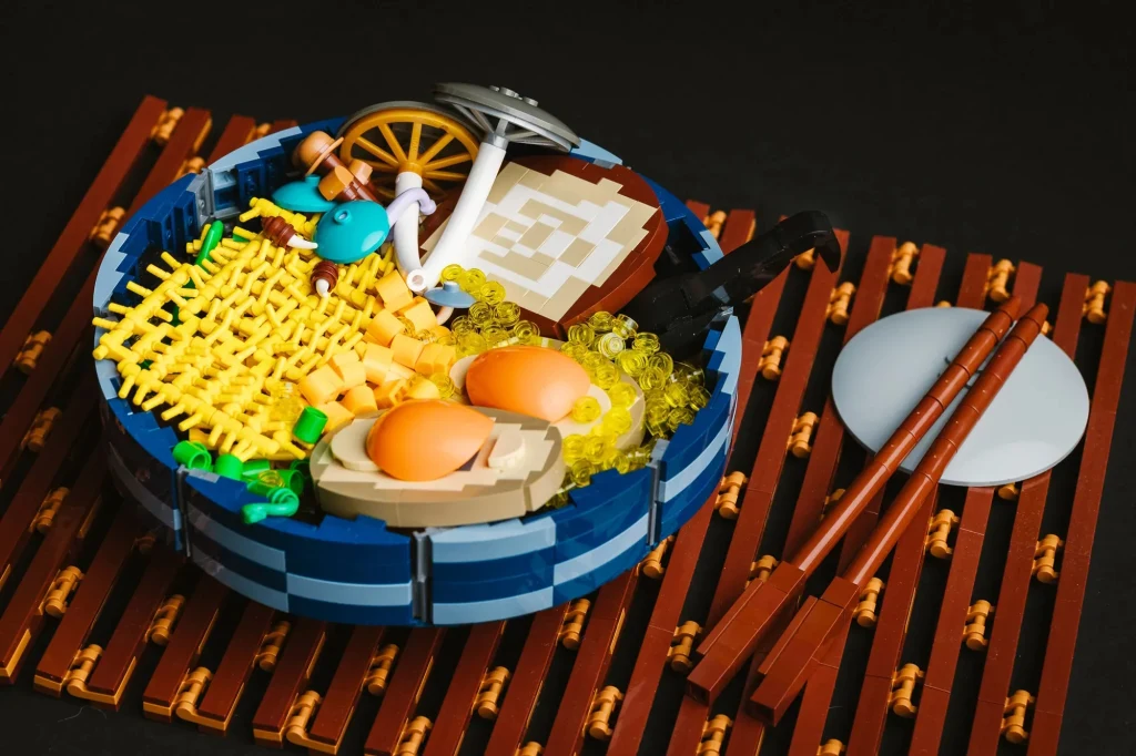 LEGO ICONS Botanicals właśnie zyskały konkurenta. Ramen z LEGO wygląda tak dobrze, że trzeba będzie uważać, by się nie zapomnieć... Miseczka ramen przedstawiona w formie kreatywnej budowli z klocków LEGO, z widocznymi charakterystycznymi składnikami jak makaron, jajka, kawałek mięsa, ozdobna płytka przypominająca narutomaki, dodatki oraz pałeczki ułożone na bambusowej podkładce.