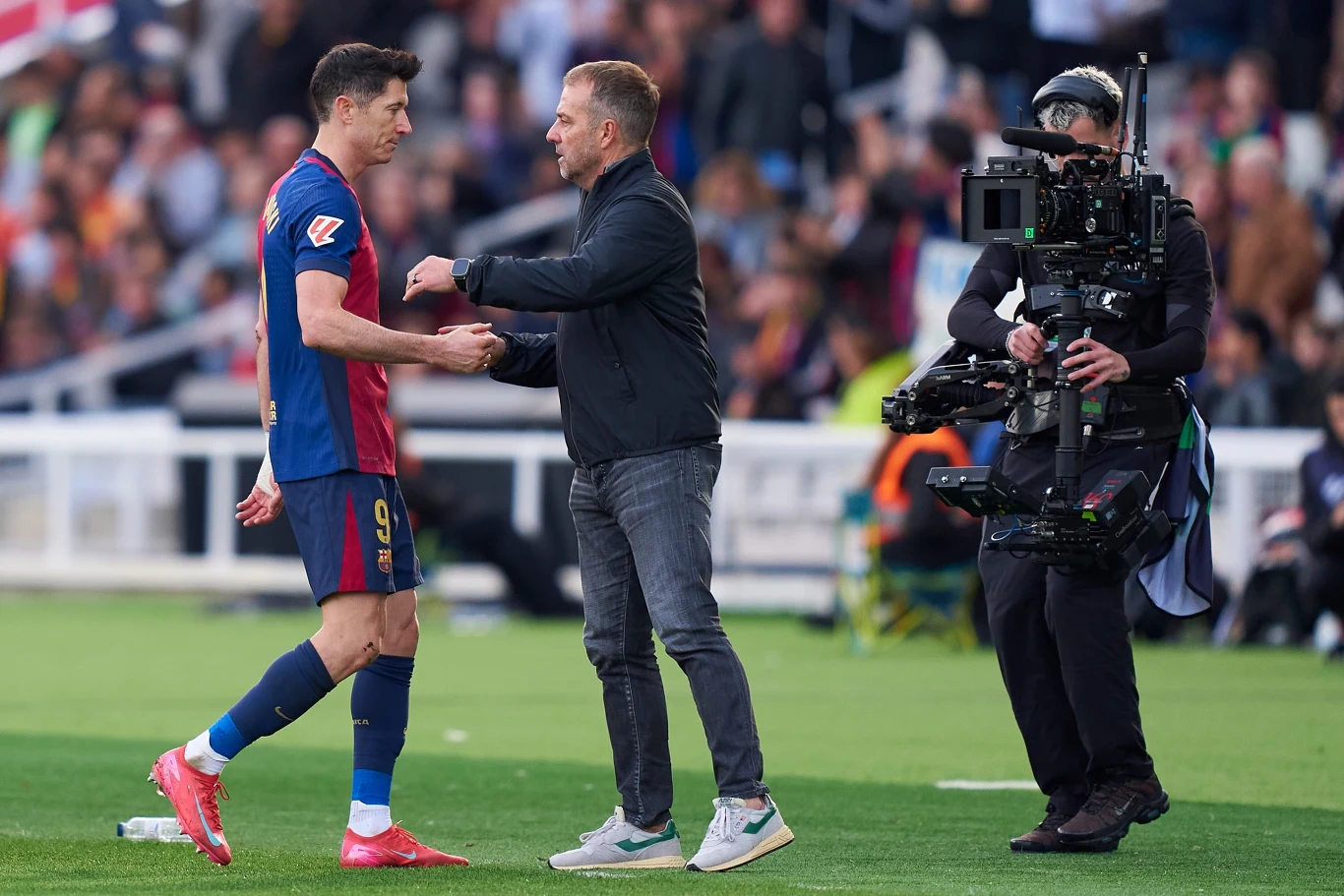 Robert Lewandowski i Hansi Flick, 02.02.2025 r. Piłkarz ubrany w koszulkę z numerem 9 w granatowo-bordowych barwach stoi na boisku, ściskając dłoń mężczyzny w casualowym ubraniu, obok kamerzysta z profesjonalną kamerą filmową, w tle zamazana publiczność.