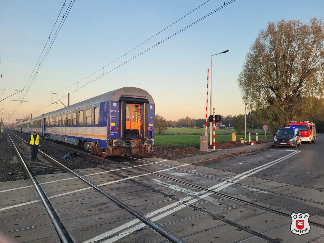 Śmiertelny wypadek w Zbąszyniu. Pociąg pasażerski zderzył się z samochodem Pociąg osobowy zatrzymany na przejeździe kolejowym przy otwartych szlabanach, w tle pole oraz pojazd służb ratunkowych i dwóch pracowników zabezpieczających miejsce.