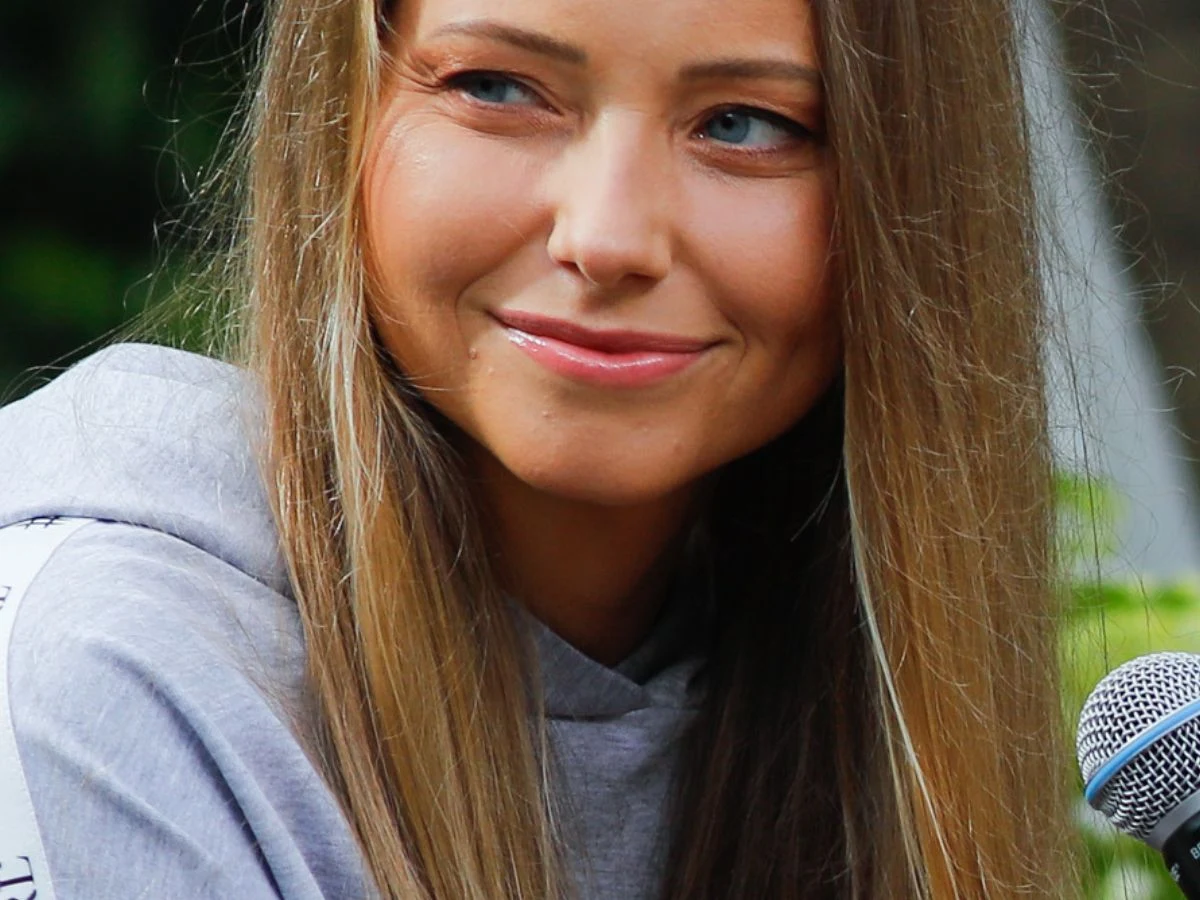 Agata Rubik Młoda kobieta z długimi, prostymi blond włosami uśmiecha się lekko, ma niebieskie oczy i ubrana jest w szarą bluzę, obok znajduje się mikrofon, tło jest rozmyte z zielenią.