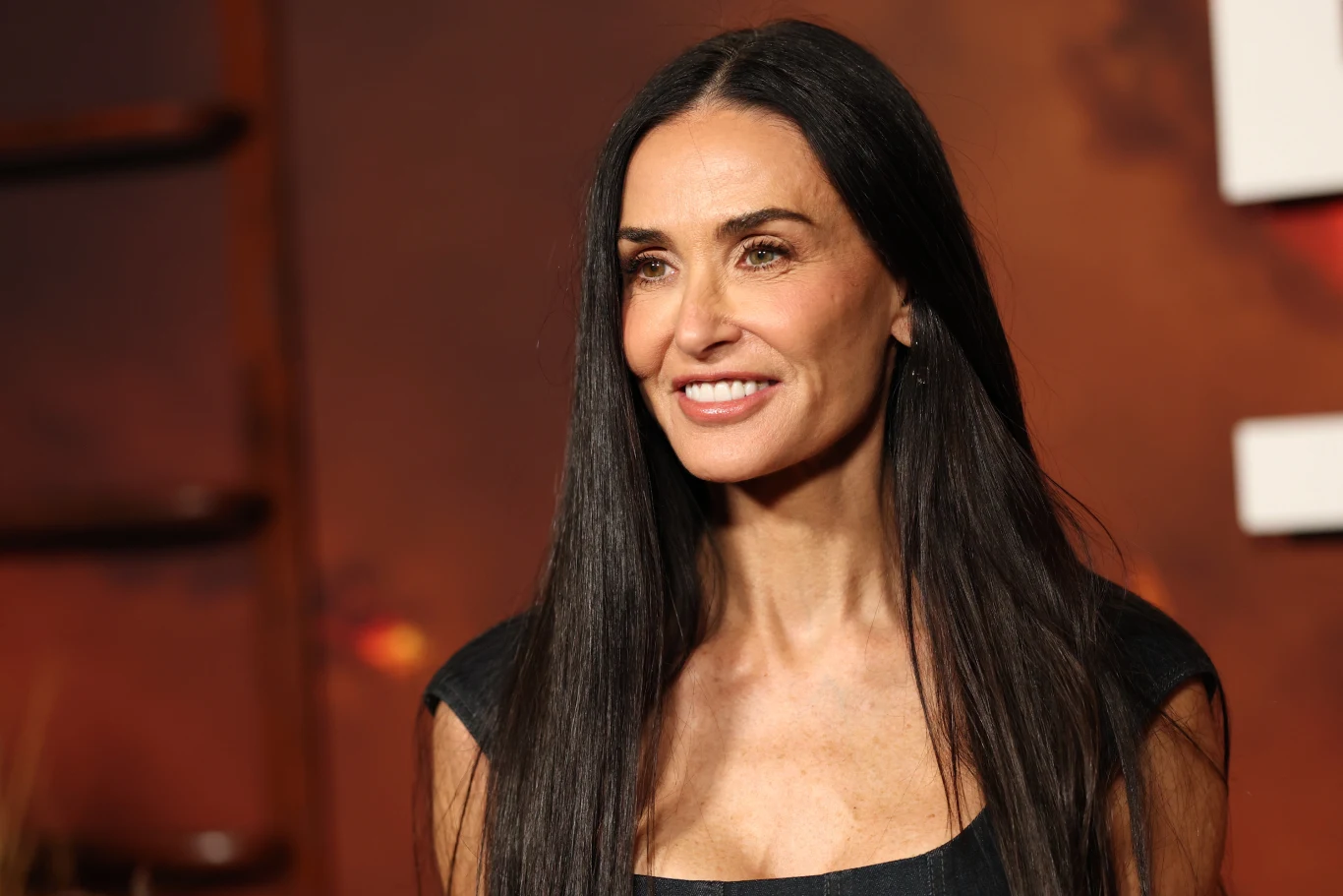 Demi Moore zachwyca coraz bardziej z każdym publicznym pojawieniem się na czerwonym dywanie Kobieta z długimi, prostymi, ciemnymi włosami ubrana w elegancką, czarną sukienkę stoi na tle rozmytego, czerwono-brązowego tła z fragmentem białego napisu po prawej stronie.