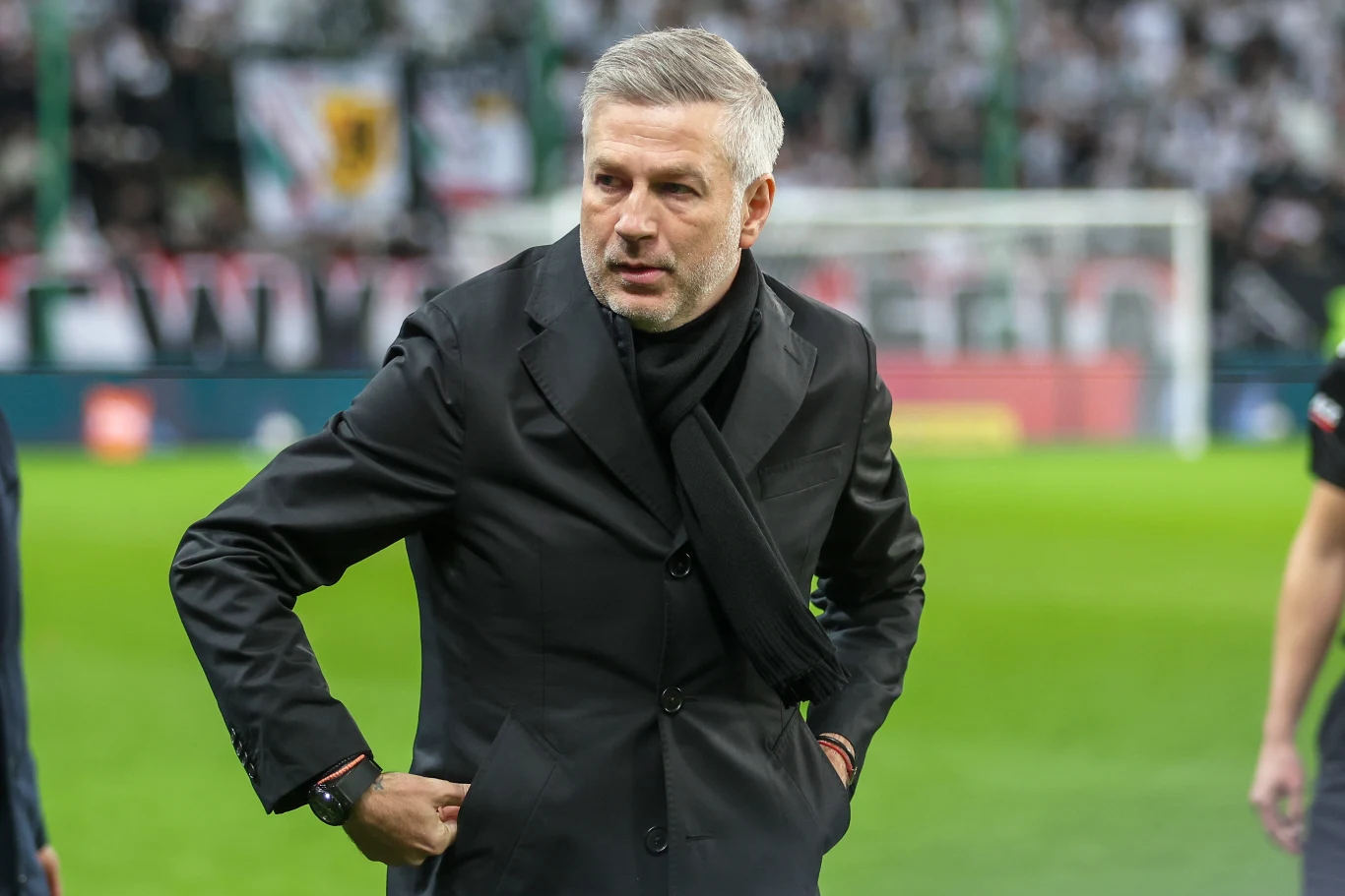 Edward Iordanescu Mężczyzna w czarnym płaszczu i szaliku stoi na tle stadionu piłkarskiego, z rękami opartymi na biodrach, skoncentrowany, w tle rozmyte trybuny i kibice.