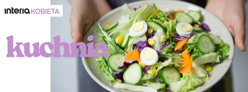 Kolorowa sałatka z plastrami ogórka, jajkiem, kukurydzą, liśćmi sałaty, marchewką i kapustą na białym talerzu trzymanym przez dwie ręce, z napisem kuchnia i logo Interia Kobieta w tle.