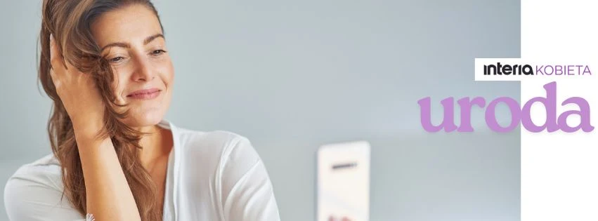 Młoda kobieta z długimi, rozpuszczonymi włosami uśmiecha się lekko, dotykając włosów ręką, w tle biała powierzchnia oraz telefon.