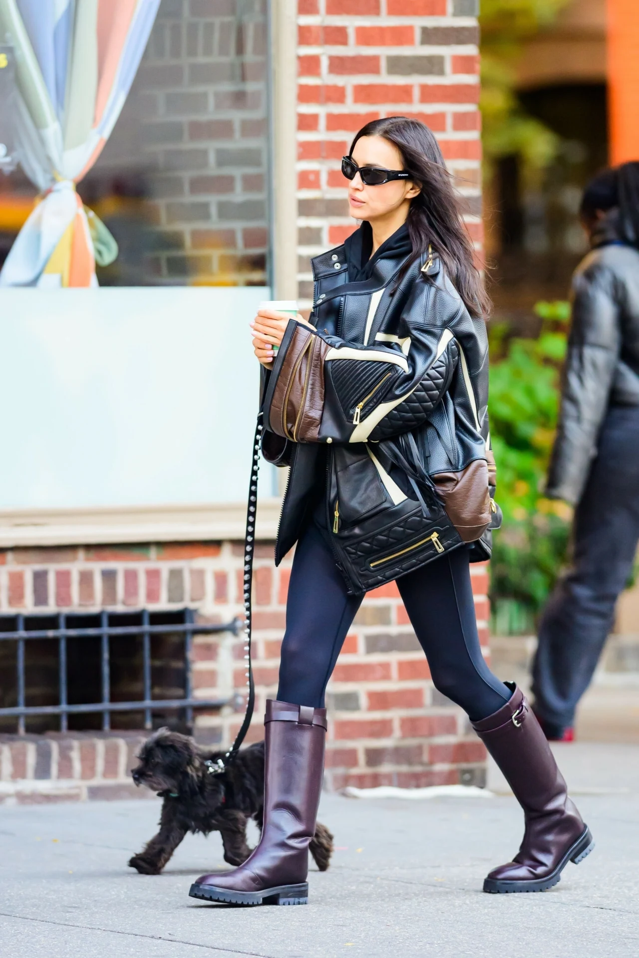 Kobieta w czarnych okularach przeciwsłonecznych, ubrana w czarną skórzaną kurtkę, legginsy i wysokie brązowe buty, spaceruje z małym czarnym psem na smyczy po chodniku w mieście, w tle fragment ceglanego budynku.