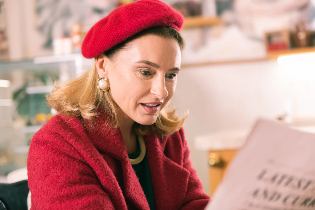 Kobieta w czerwonym berecie i płaszczu siedzi w kawiarni i czyta gazetę, koncentrując się na tekście. W tle widoczna rozmyta półka z przedmiotami codziennego użytku.