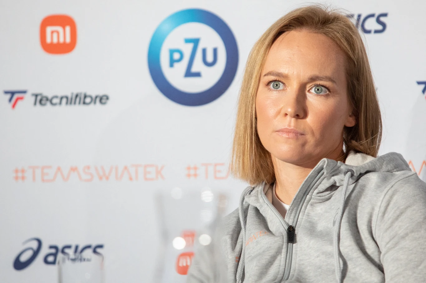 Daria Abramowicz Kobieta o jasnych włosach w sportowej bluzie, siedzi na tle tablicy z logotypami sponsorów takich jak PZU, Asics, Tecnifibre oraz Xiaomi.