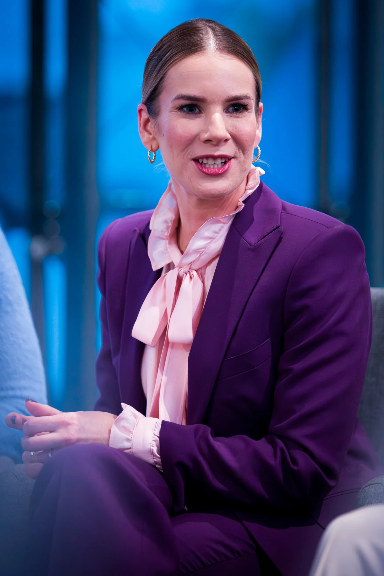 Kobieta w eleganckim fioletowym garniturze i jasnoróżowej bluzce z kokardą, siedząca w studiu z niebieskim tłem, uśmiechnięta, z zadbanymi włosami spiętymi w gładki kucyk, nosi złote kolczyki.