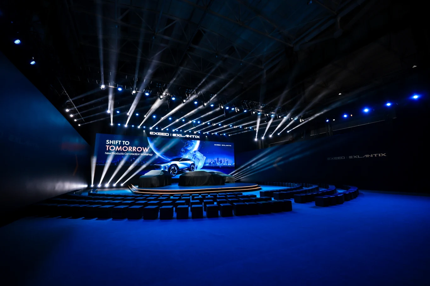 Pierwsze spotkania z elektrycznymi samochodami marki Exlantix zaostrzyły apetyt kierowców Nowoczesna scena oświetlona licznymi reflektorami, na której znajduje się samochód sportowy na tle dużego ekranu z napisem 'Shift to tomorrow', wokół ustawione są rzędy pustych siedzeń tworzących półokrąg.