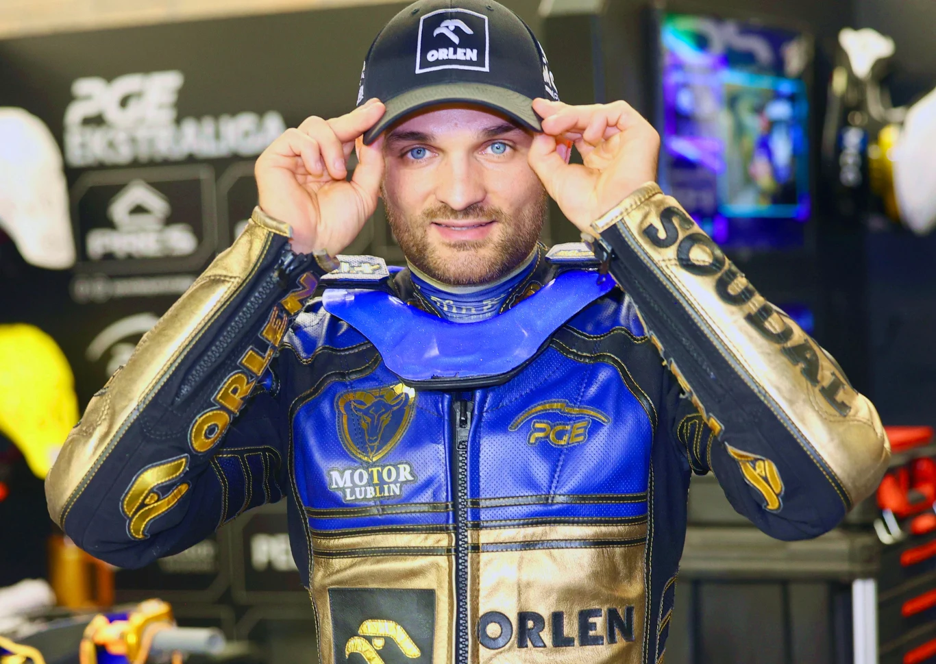 Mężczyzna w złoto-niebieskim kombinezonie żużlowym i czapce z logo Orlen w otoczeniu sprzętu sportowego i reklamowego na tle ekspozycji sponsorskiej.