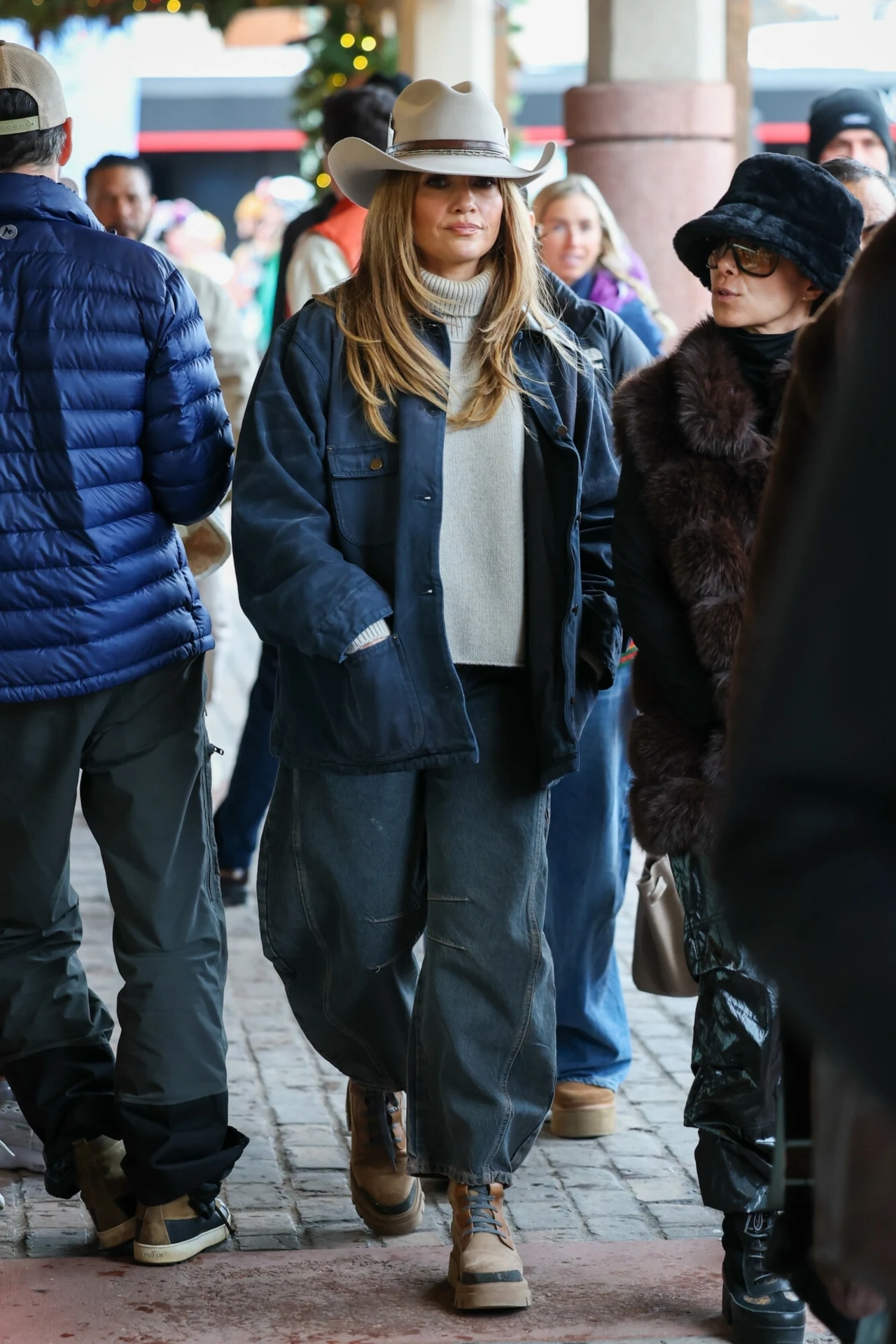 Jennifer Lopez coraz częściej sięga po kapelusze Kobieta w jasnym kapeluszu i ciemnych, luźnych ubraniach spaceruje w tłumie ludzi na ulicy. W tle widać osoby ubrane na zimowo, niektóre noszą czapki i futra. Kobieta znajduje się w centrum kompozycji, patrzy na wprost.