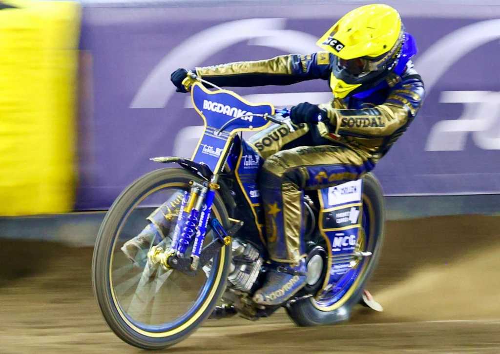 Motocyklista ubrany w żółty kask i kombinezon startuje na żużlowym torze, jadąc na motocyklu wyścigowym z widocznymi sponsorami i dynamiczną postawą w zakręcie.