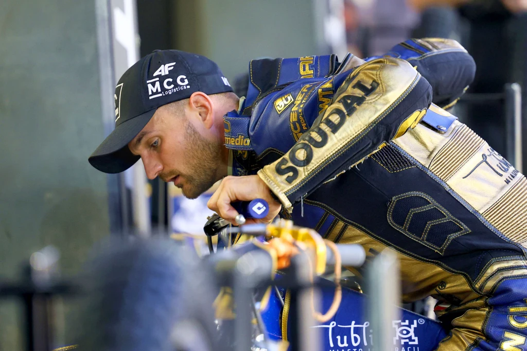 Zawodnik w stroju sportowym z logotypami sponsorów pochylony nad motocyklem żużlowym, koncentrujący się na przygotowaniu maszyny do wyścigu.