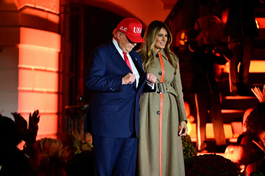 Melania Trump często wybiera proste, eleganckie stylizacje Mężczyzna w garniturze i czerwonej czapce z napisem 'USA' stoi obok kobiety w jasnym płaszczu z pomarańczowymi akcentami, oboje uśmiechnięci, w tle widoczne dekoracje w jesiennej kolorystyce i oświetlenie.