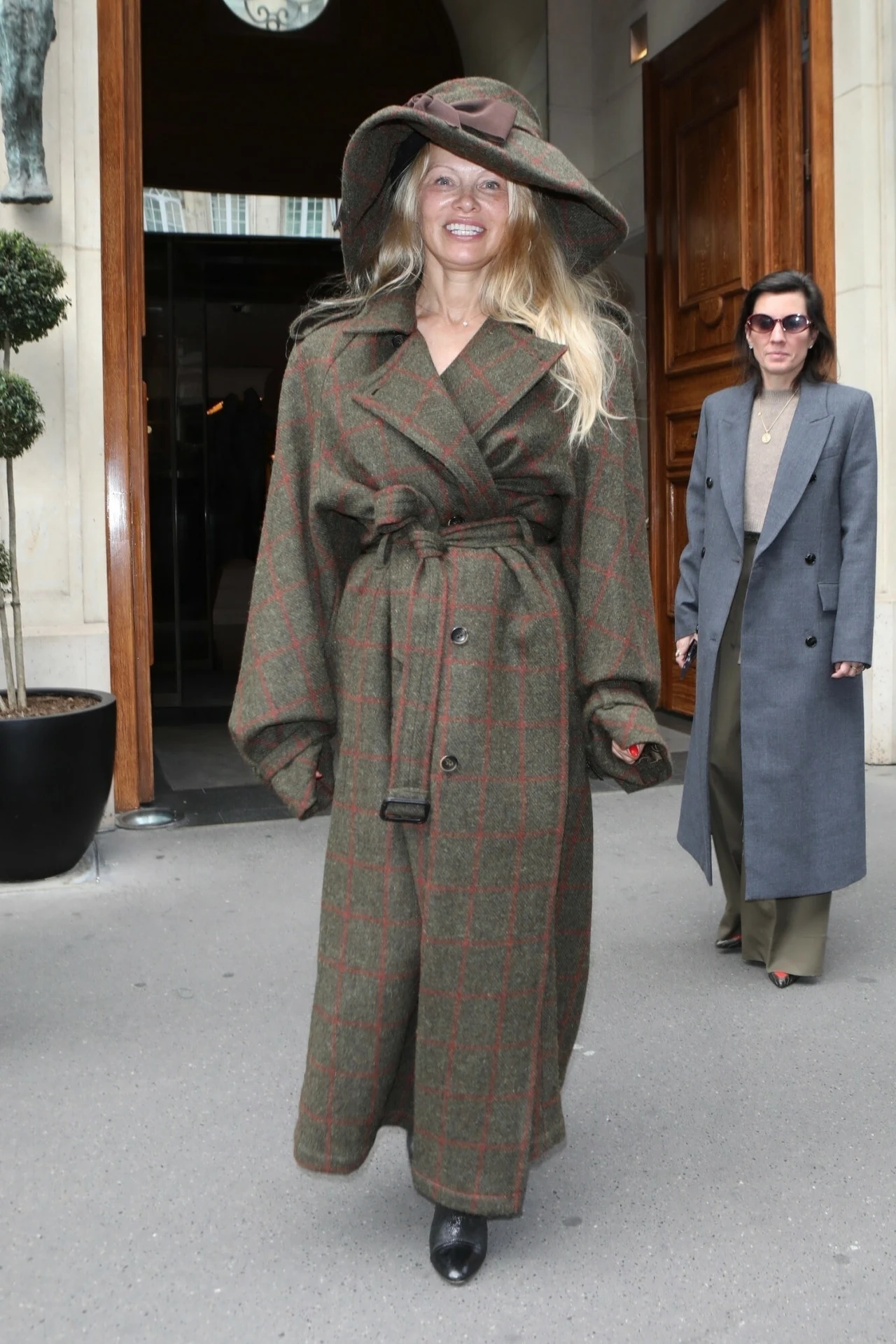 Pamela Anderson w kapeluszu imponujących rozmiarów Kobieta w szerokim płaszczu o długich rękawach i kapeluszu z szerokim rondem stoi przed drzwiami, w tle druga kobieta ubrana w szary garnitur i okulary przeciwsłoneczne.