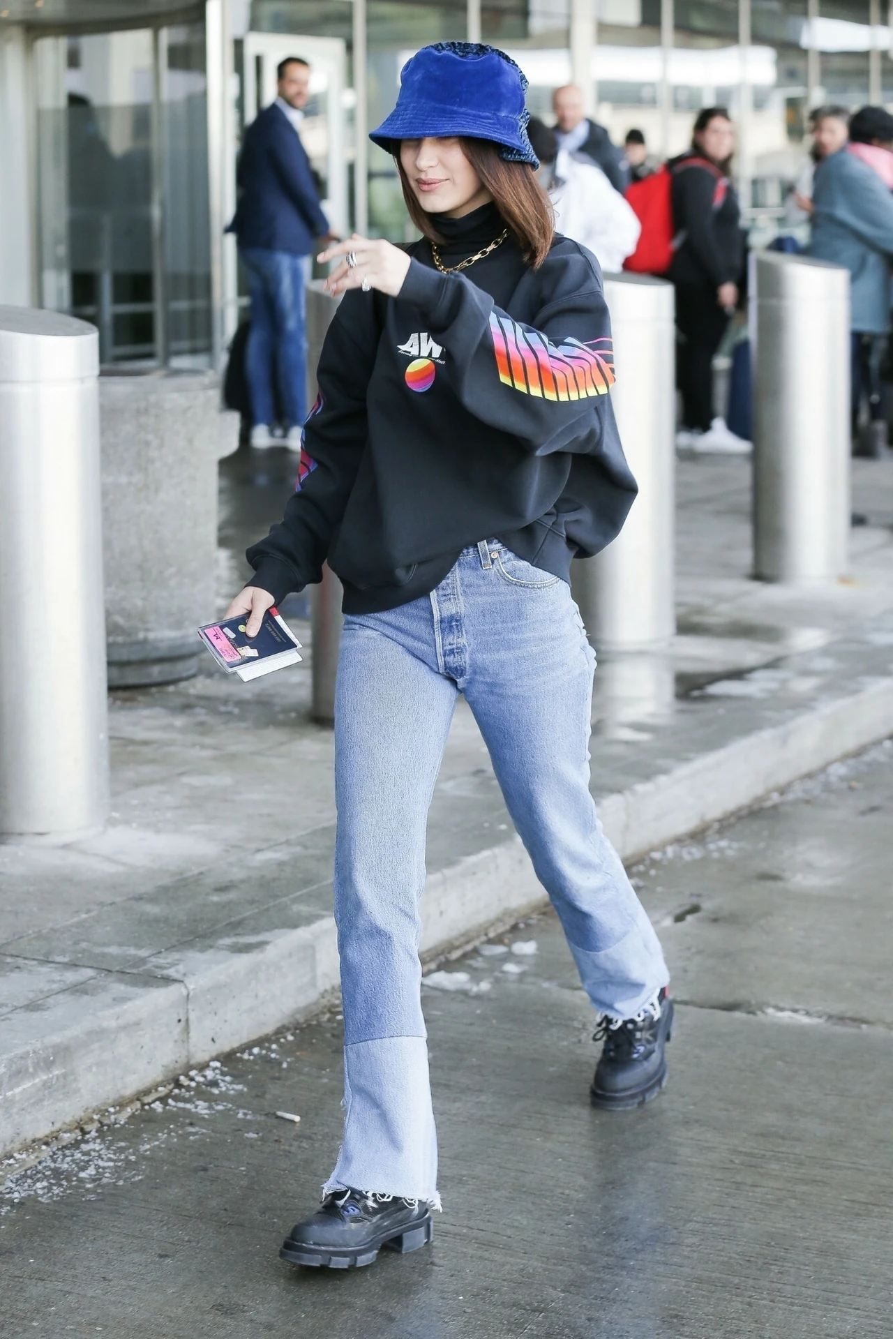Bella Hadid w niebieskim bucket hat Kobieta ubrana w czarną bluzę z kolorowymi wzorami na rękawach, niebieskie jeansy i granatowy kapelusz typu bucket hat idzie po chodniku w pobliżu wejścia do nowoczesnego budynku, wokół widocznych jest kilku przechodniów.