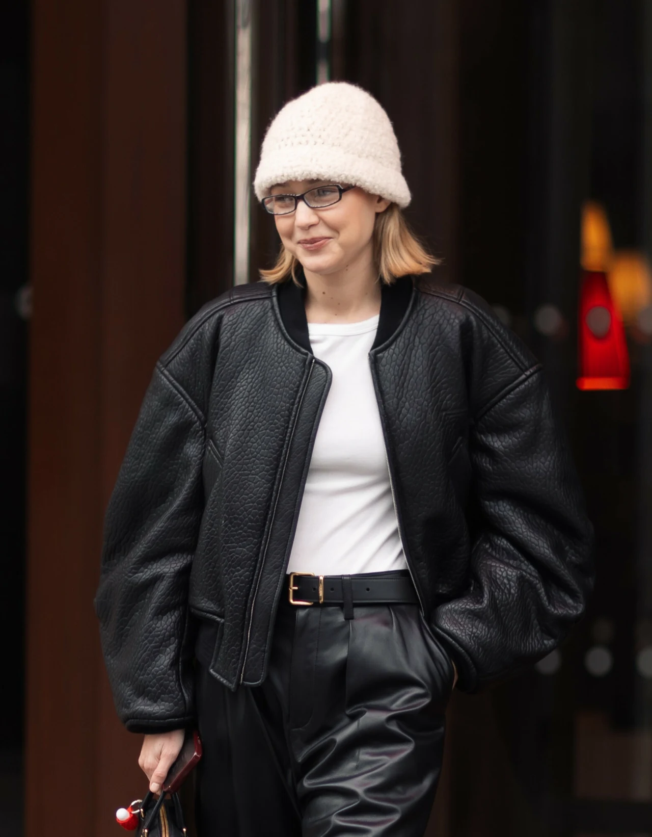 Gigi Hadid w białej czapce w stylu bucket hat Kobieta w białej dzianinowej czapce, okularach, czarnej skórzanej kurtce i spodniach, z białą koszulką, trzymająca torebkę, spacerująca po mieście.
