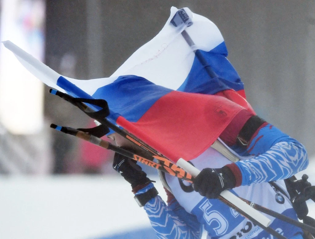 Rosjanie są wściekli po wykluczeniu przez FIS Zawodnik ubrany w sportowy strój narciarski niesie dużą rosyjską flagę, która zasłania jego twarz. W rękach trzyma kijki narciarskie. Tło rozmyte, skupia uwagę na sportowcu i fladze.