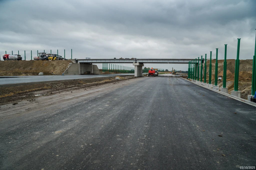 Nowa droga asfaltowa w budowie z wiaduktem przebiegającym nad jezdnią, po obu stronach montowane są zielone barierki dźwiękochłonne, widoczne maszyny budowlane oraz chmurne niebo.