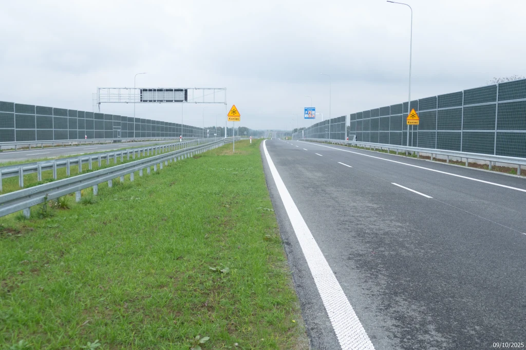 Nowoczesna, szeroka autostrada oddzielona barierkami ochronnymi i ekranami dźwiękochłonnymi, otoczona pasem zieleni, na jezdni brak pojazdów, widoczne znaki drogowe oraz tablice informacyjne przy pochmurnym niebie.