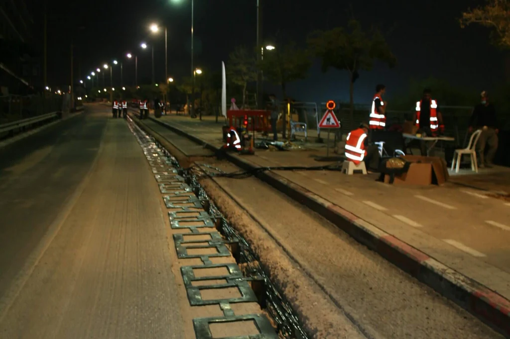 Nocna praca drogowa przy linii tramwajowej, kilku pracowników w odblaskowych kamizelkach wykonuje prace serwisowe, ulica jest oświetlona latarniami i wyłączona z ruchu, widoczny sprzęt do prac drogowych.