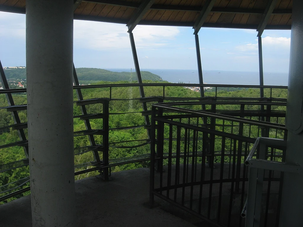Wieża widokowa w Kolibkach zapewnia widoki na Bałtyk - konkretnie na Zatokę Gdańską. Taras widokowy ze schodami i balustradą otoczony zielonym lasem, w tle rozciąga się panorama wzgórz i morza pod niebieskim niebem.