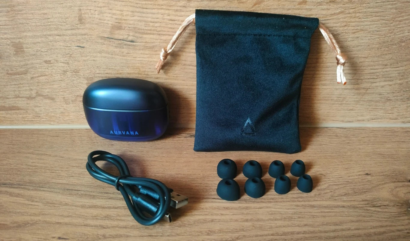 Etui z napisem Aurvana, czarny woreczek z bawełnianym sznurkiem, sześć sylikonowych końcówek do słuchawek oraz kabel USB, wszystko ułożone na drewnianej powierzchni.