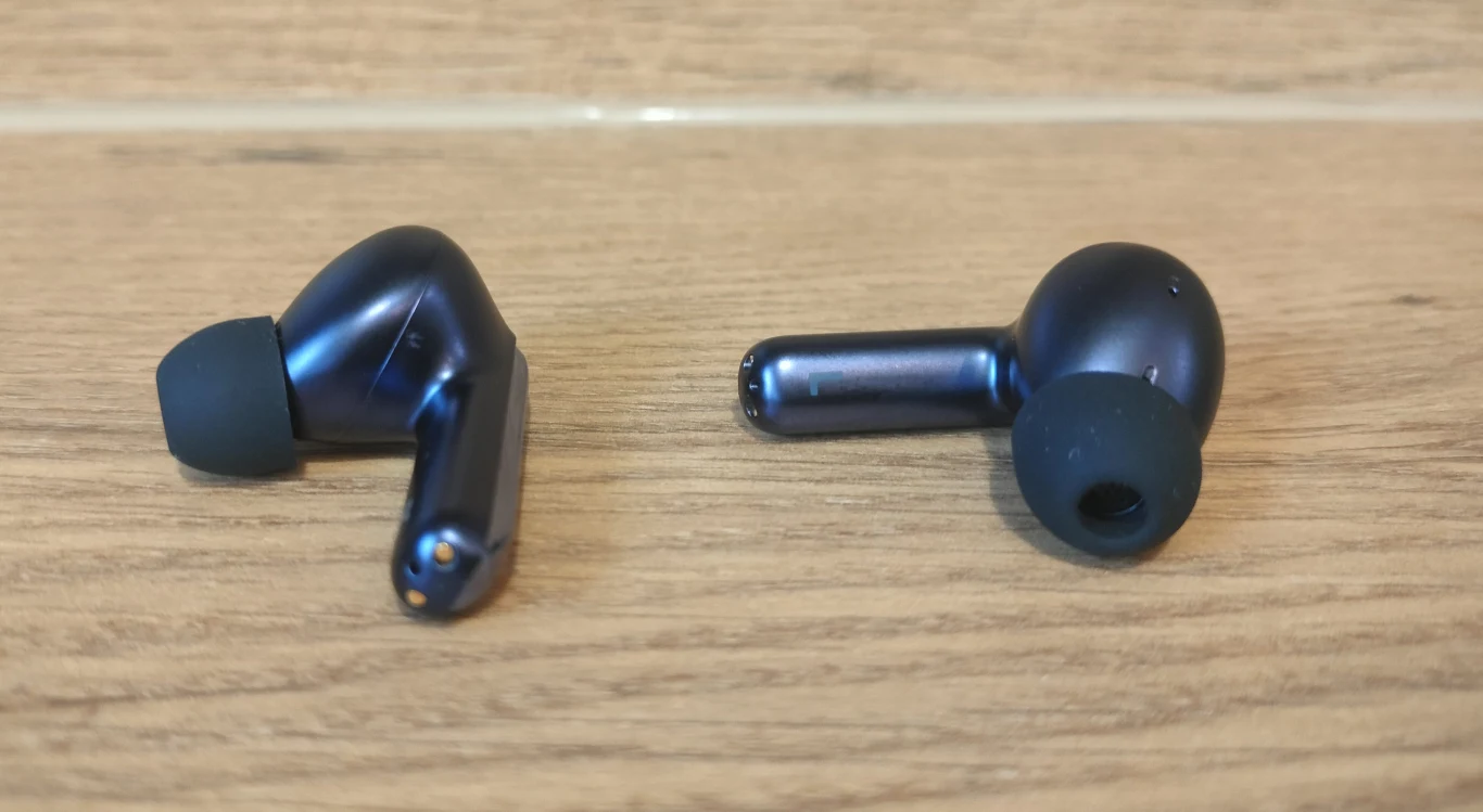 Bezprzewodowe słuchawki douszne o ciemnej metalicznej obudowie i silikonowych nakładkach leżące na drewnianej powierzchni.