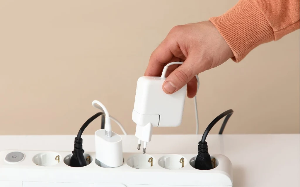 Czy podłączona ładowarka pobiera energię? Ręka w pomarańczowym rękawie podłącza biały zasilacz do przedłużacza, w którym znajdują się już inne wtyczki elektryczne.