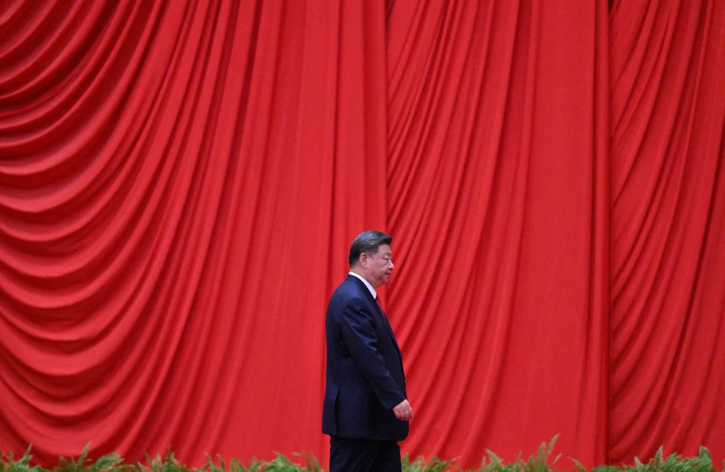 Mężczyzna ubrany w ciemny garnitur idzie wzdłuż bogato marszczonej czerwonej kotary, na dole widoczne zielone rośliny. Dominującym elementem jest intensywnie czerwona tkanina tworząca tło.