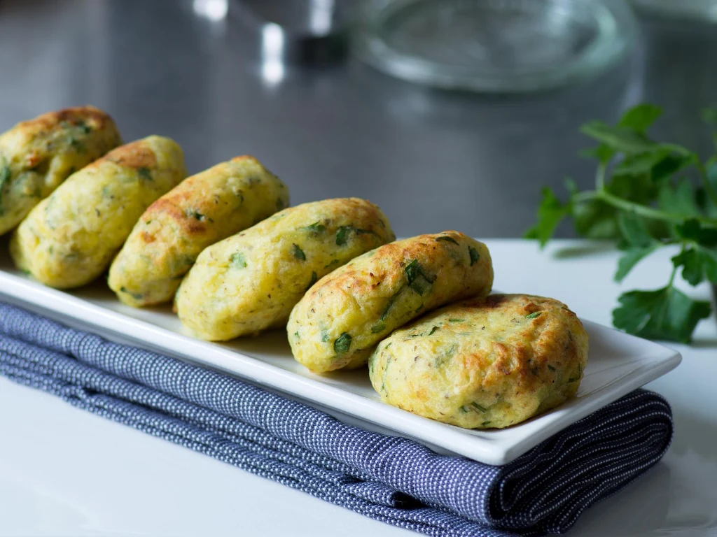 Ziemniaki pozostałe po obiedzie mogą posłużyć do zrobienia kotlecików Rząd pięciu upieczonych kotletów ziemniaczanych z ziołami ułożonych na podłużnym, białym talerzu na ciemnoniebieskiej serwetce, w tle delikatnie rozmyte świeże zioła.
