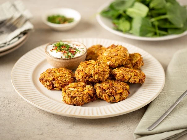 Z obierek powstaną aromatyczne, chrupiące kotleciki Talerz z kilkoma złocistymi placuszkami warzywnymi podanymi z sosem jogurtowym w małej miseczce, obok widoczne świeże liście szpinaku, szara serwetka i widelec.