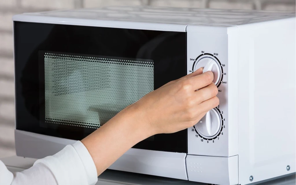Twoja mikrofalówka ma wyświetlacz LED? Sprawdź, czy nie pochłania więcej prądu niż powinna Ręka osoby nastawia pokrętła na białej kuchence mikrofalowej ustawionej na blacie, z widoczną przednią szybą i panelami sterowania.