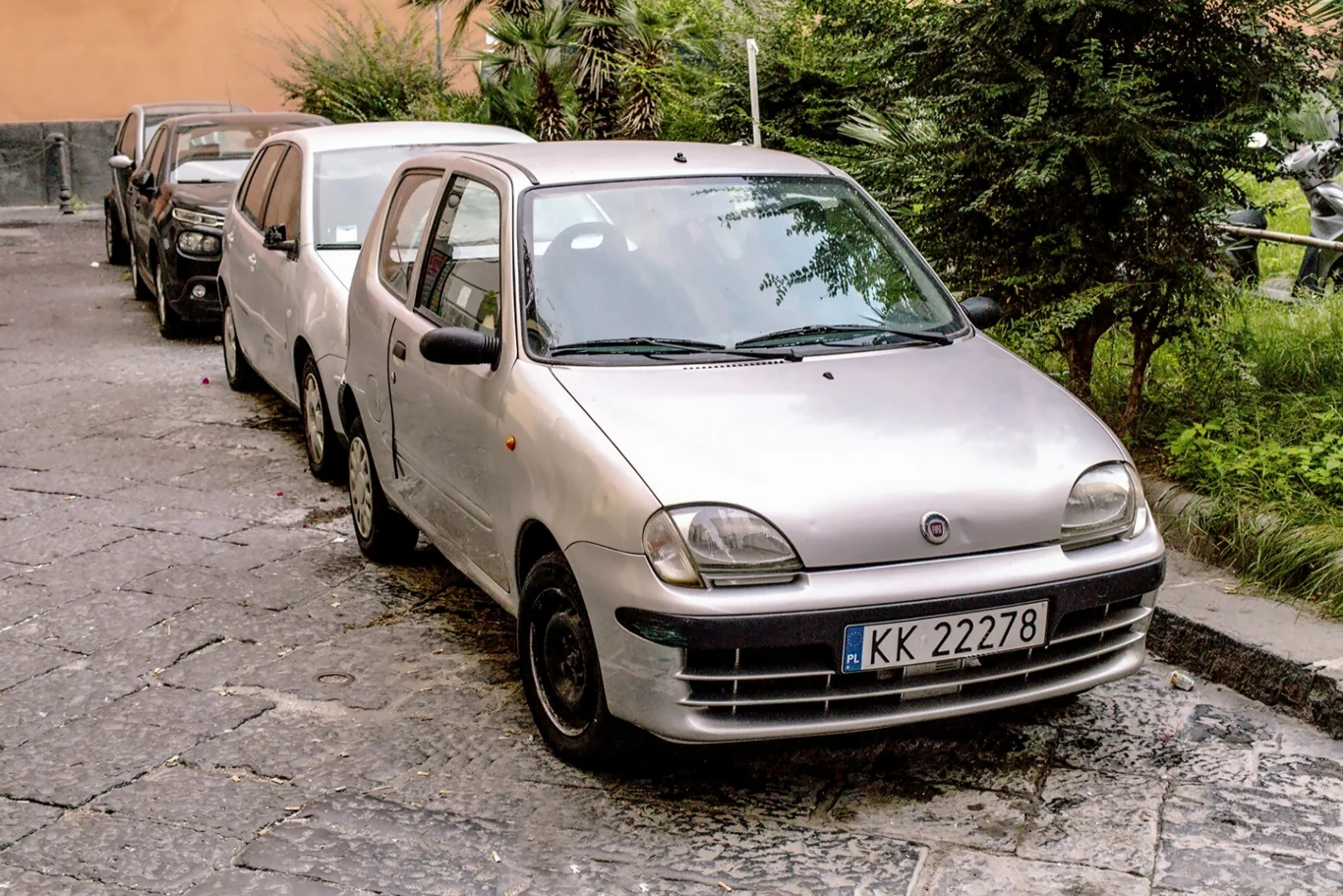 Auta na polskich rejestracjach we Włoszech. O co chodzi? Cztery samochody zaparkowane na wąskiej ulicy, na pierwszym planie srebrny fiat z tablicą rejestracyjną KK 22278, wokół roślinność i budynek o pomarańczowej elewacji w tle.