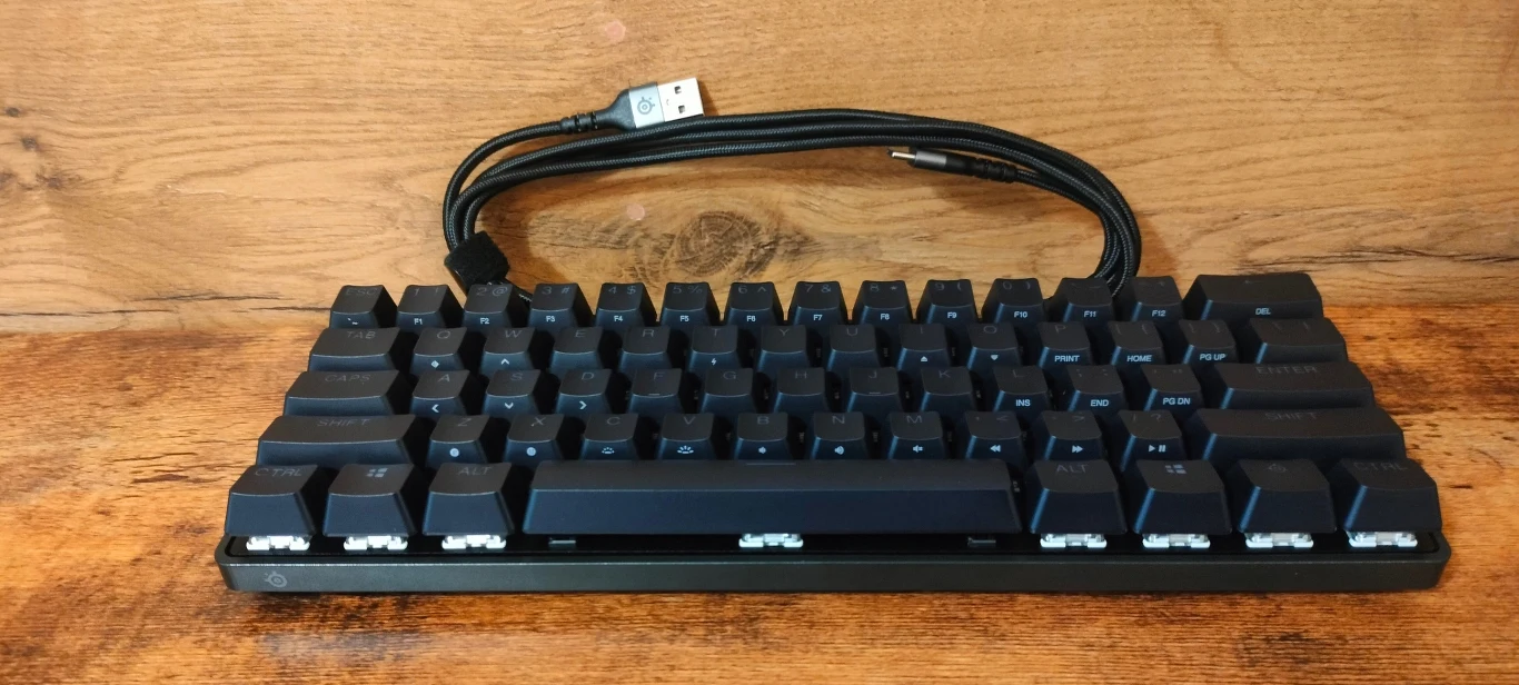 Czarna, mechaniczna klawiatura komputerowa bez klawiszy numerycznych, z dołączonym czarnym kablem USB, ustawiona na drewnianej powierzchni, w tle drewniana ściana.