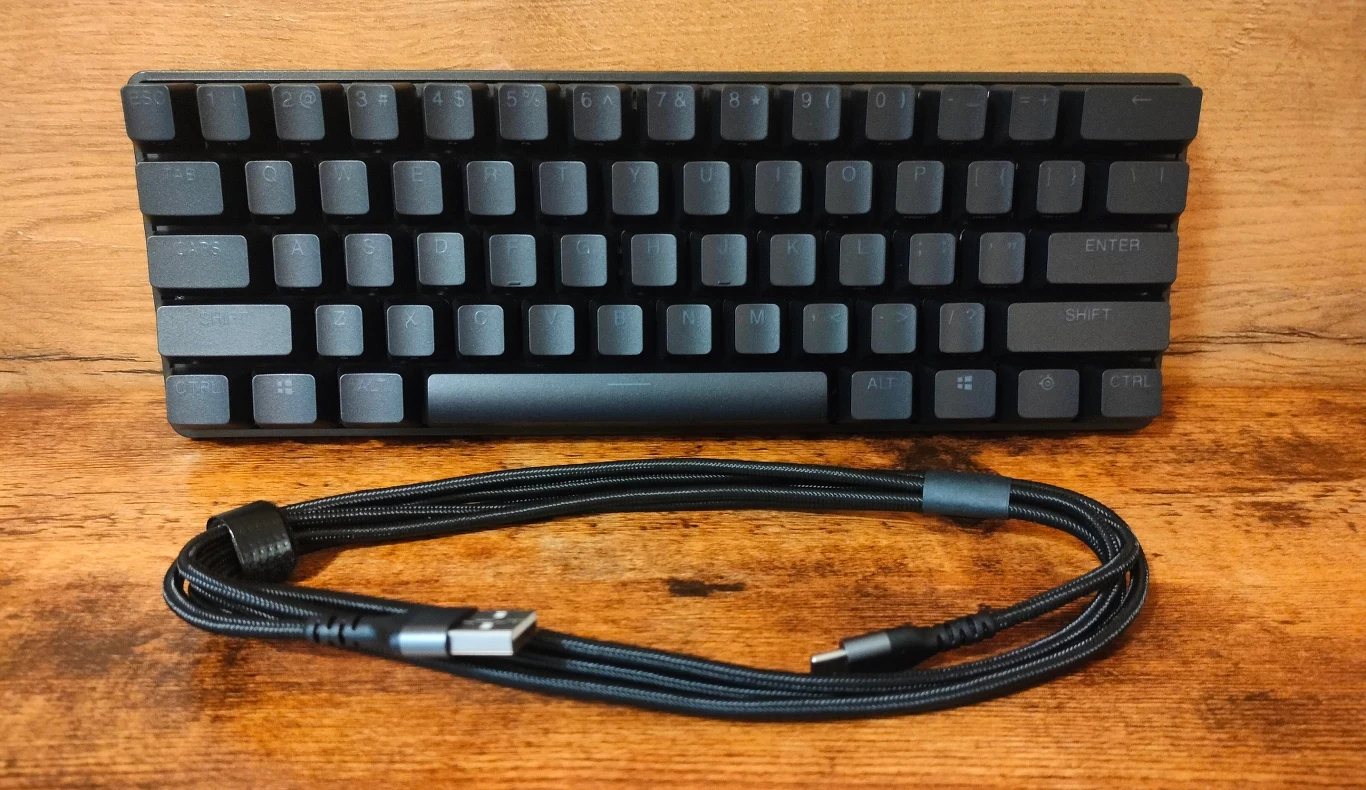 Czarna, kompaktowa klawiatura mechaniczna z minimalistycznym układem klawiszy, ustawiona na drewnianym tle, poniżej widoczny czarny przewód USB typu C z oplotem zapewniającym dodatkową wytrzymałość.