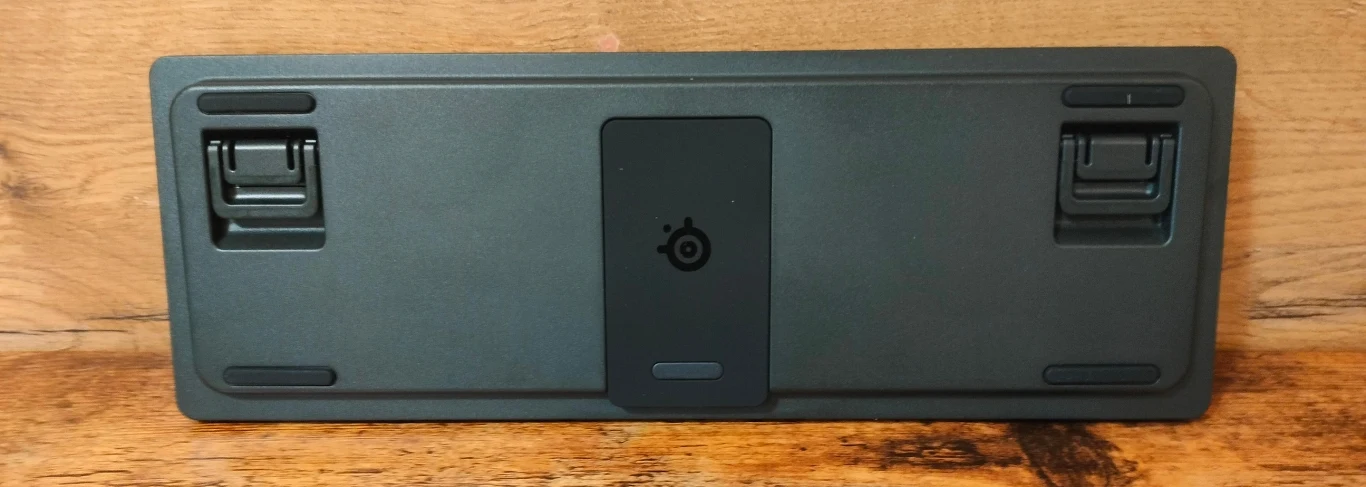 Czarna, prostokątna podkładka pod klawiaturę umieszczona na drewnianej powierzchni z widocznym logo producenta na środku oraz dwoma składanymi nóżkami po bokach.