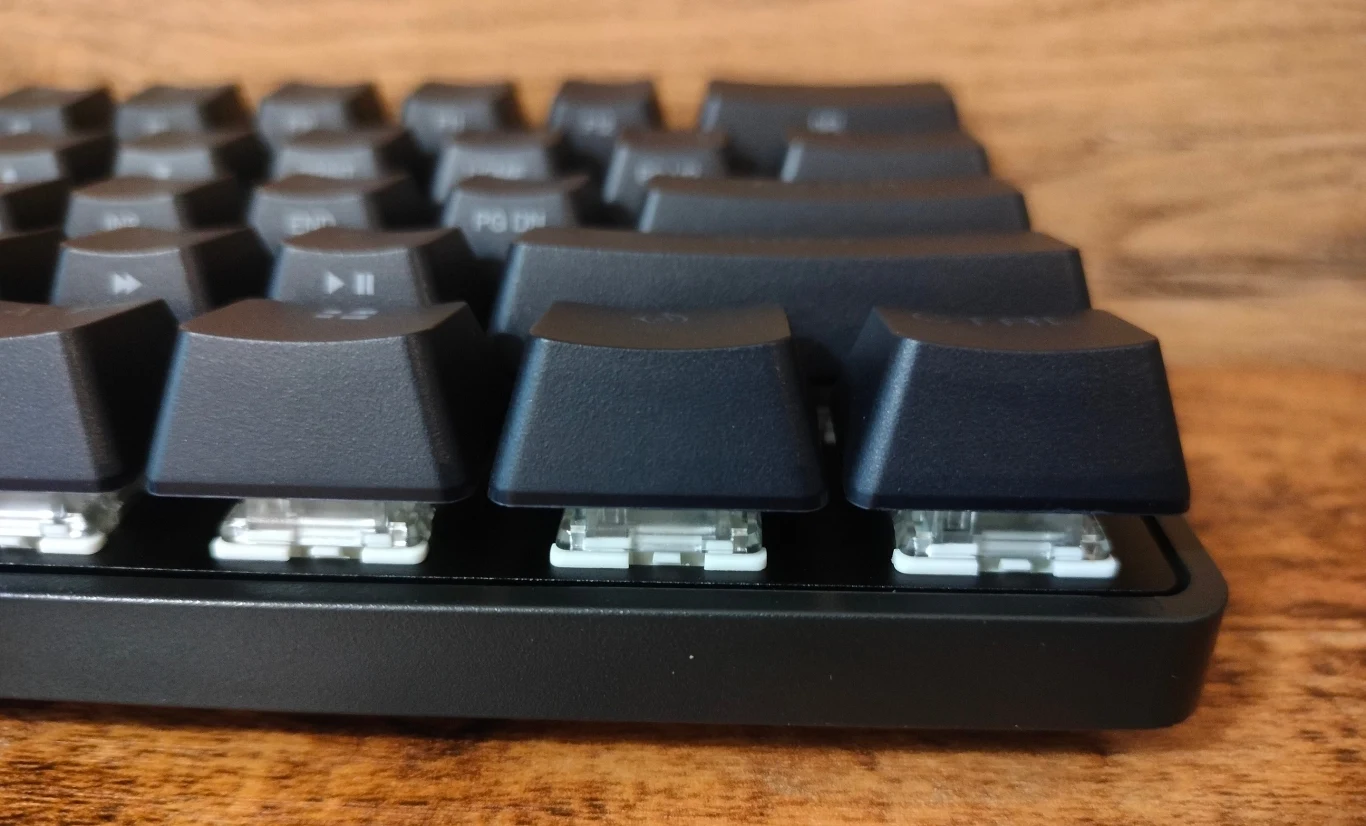 Część mechanicznej klawiatury komputerowej z widocznymi klawiszami i przezroczystymi przełącznikami, tło z jasnego drewna.