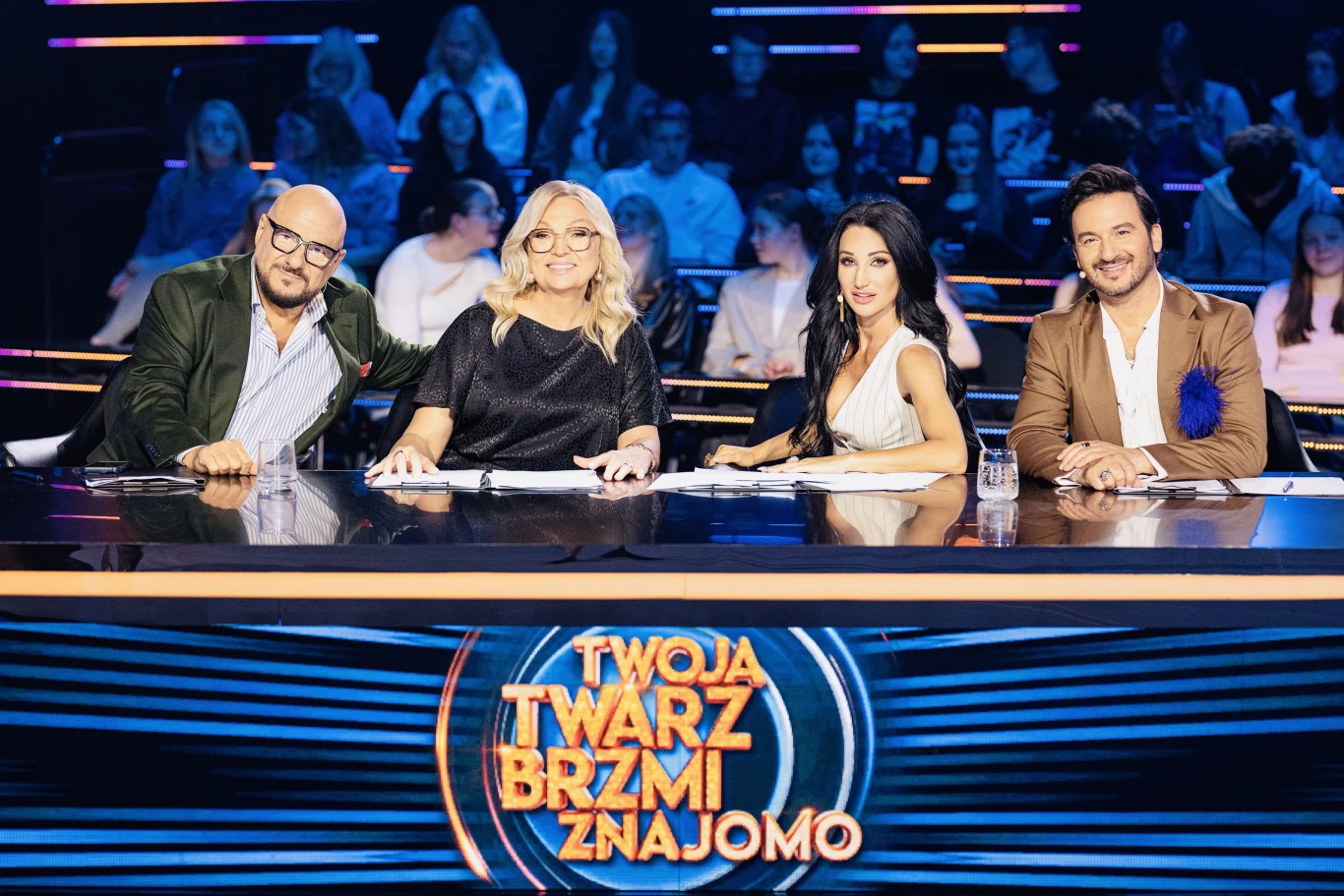 Jury programu "Twoja twarz brzmi znajomo": Piotr Gąsowski, Małgorzata Walewska, Justyna Steczkowska i Stefano Terrazzino Czworo jurorów siedzi przy stole jurorskim na planie programu rozrywkowego, za nimi widownia, na pierwszym planie logo programu "Twoja Twarz Brzmi Znajomo".