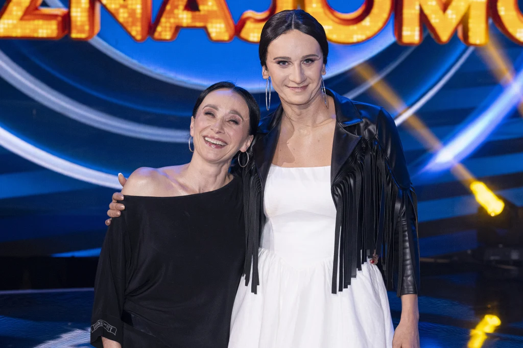 Renata Przemyk i Modest Ruciński (jako Renata Przemyk) w "Twoja twarz brzmi znajomo" Dwie uśmiechnięte kobiety pozują razem na tle kolorowego, scenicznego napisu. Jedna z nich ubrana jest w czarną bluzkę, druga ma na sobie białą sukienkę i czarną kurtkę z frędzlami. Oświetlenie sceniczne podkreśla atmosferę wydarzenia.