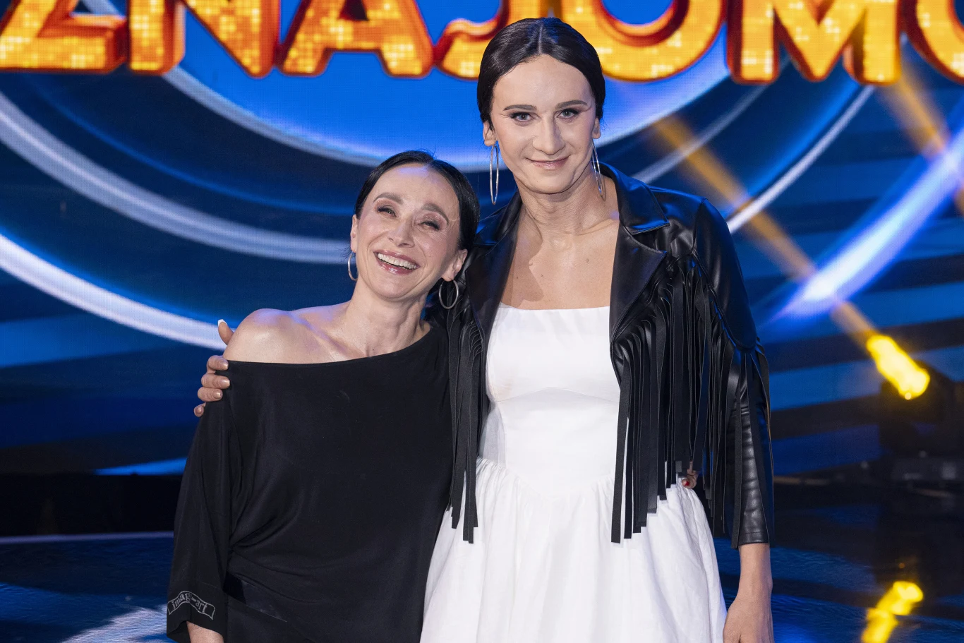 Renata Przemyk i Modest Ruciński (jako Renata Przemyk) w "Twoja twarz brzmi znajomo" Dwie uśmiechnięte kobiety pozują razem na tle kolorowego, scenicznego napisu. Jedna z nich ubrana jest w czarną bluzkę, druga ma na sobie białą sukienkę i czarną kurtkę z frędzlami. Oświetlenie sceniczne podkreśla atmosferę wydarzenia.