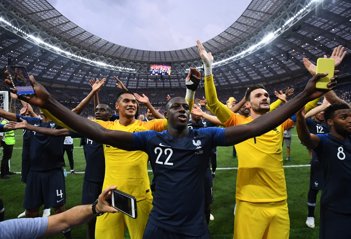 Benjamin Mendy, mistrz świata z 2018 roku, niebawem powinien zagrać w barwach... drugiego zespołu Pogoni Szczecin Piłkarze drużyny w niebieskich i żółtych strojach świętują zwycięstwo na stadionie, unosząc ręce w geście radości, wokół nich kibice wykonują zdjęcia smartfonami, a tło wypełnia ogromna, zadaszona trybuna.