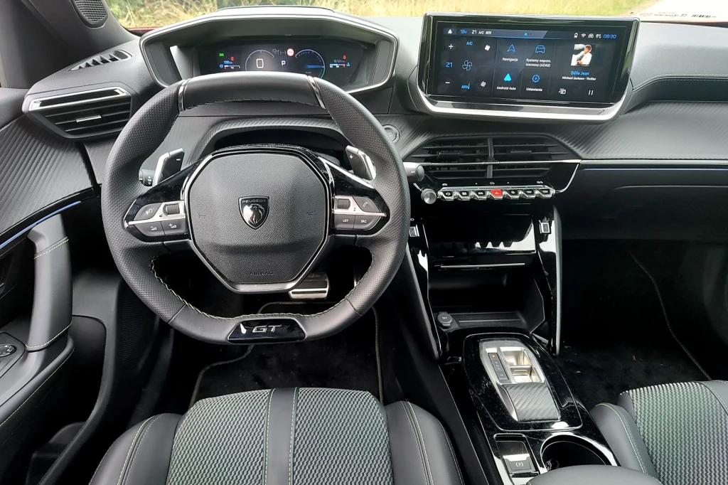 Na ekran cyfrowych zegarów patrzymy nie przez, ale ponad wieńcem małej kierownicy Centralna część wnętrza samochodu z nowoczesnym kierownicą, cyfrowym zestawem wskaźników oraz dużym ekranem multimedialnym. Konsola środkowa wyposażona w przyciski i selektor zmiany biegów. Fotele wykonane z materiałów wysokiej jakości.